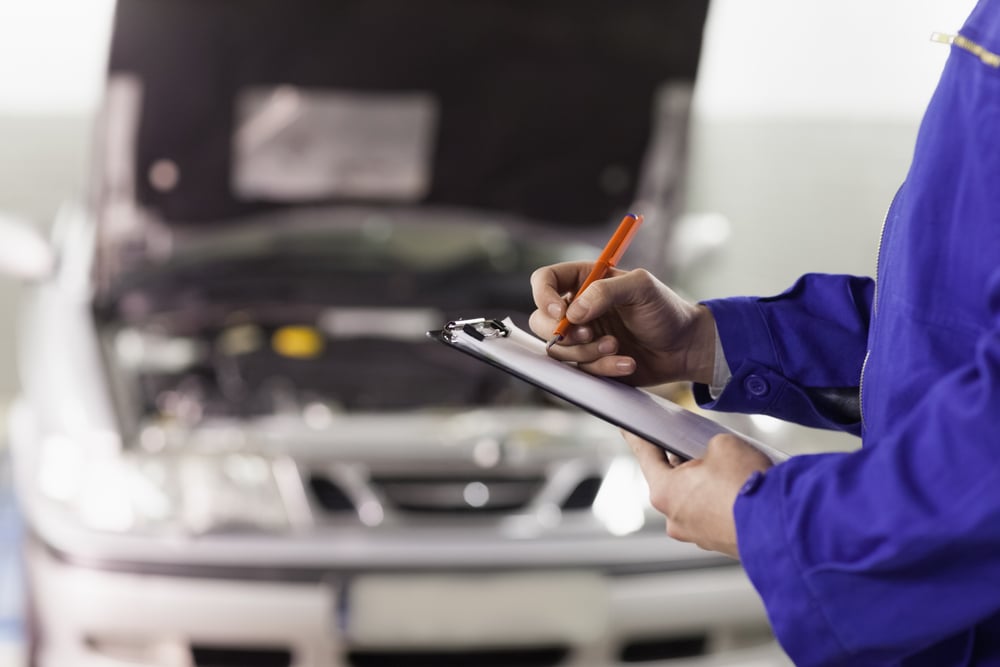 mechanic holding a notepad and standing in front of a car with hood open 